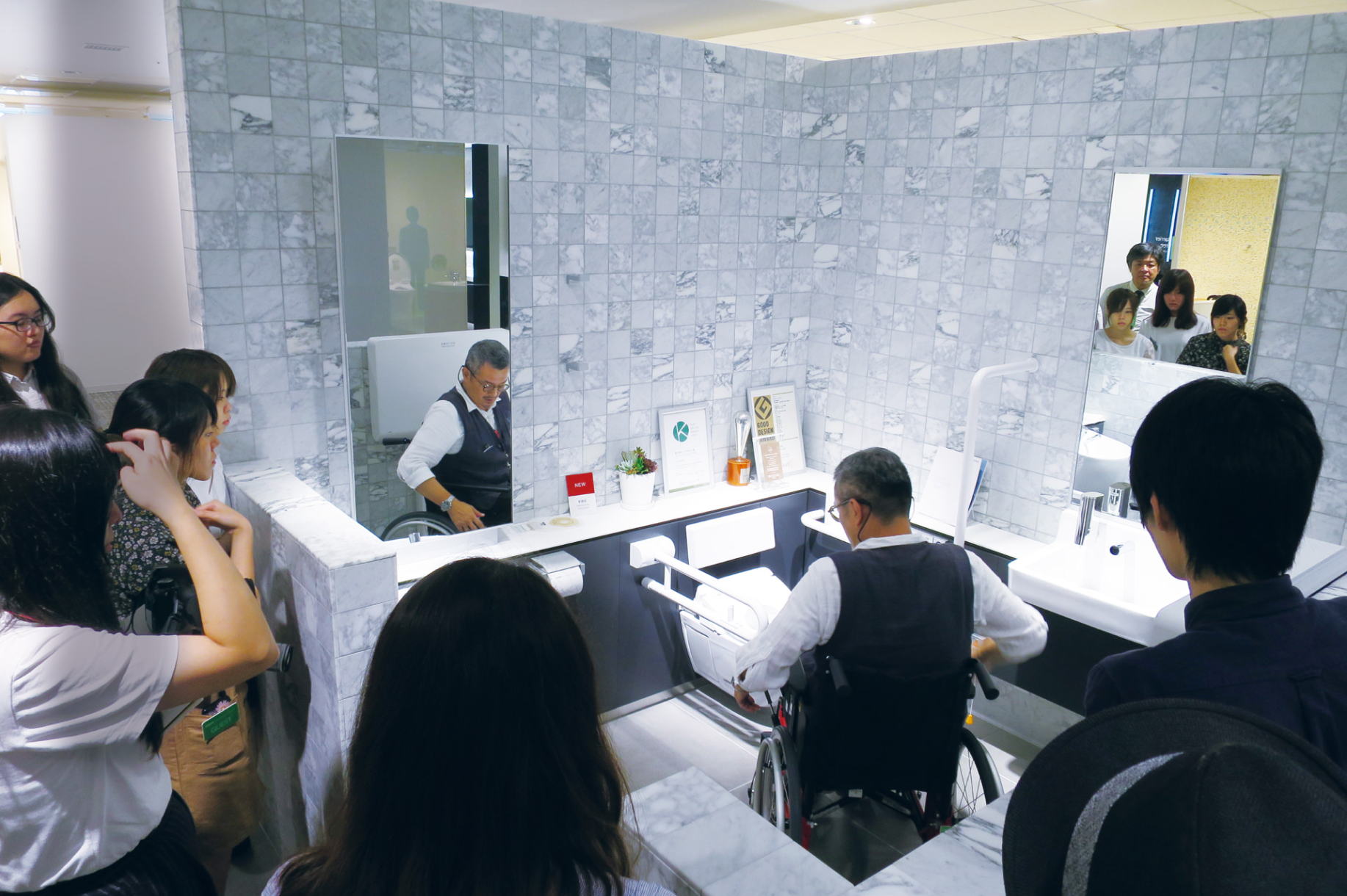 洗面所で車椅子を使用する人向けの設備を紹介している展示会の様子。壁と洗面台はタイル調で、鏡には参加者の姿が映っている。